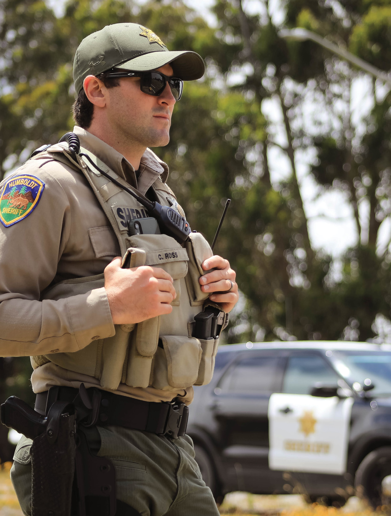 Sheriff's deputy standing with a patrol car behind him.