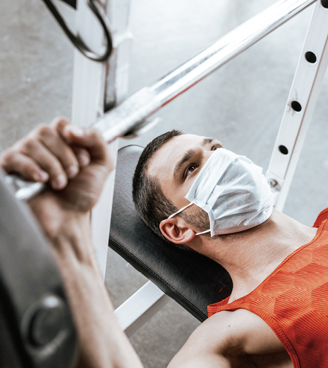 Selective focus of sportsman in medical mask exercising with barbell in gym.