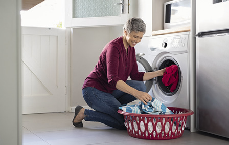 Happy senior woman loading dirty clothes in washing machine. Smiling mature woman sitting on floor putting clothed in washing machine from laundry basket. Housework.