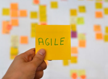 A hand is holding a orange agile sticker and there is a Kanban board of agile methodology on the background, which is a developing trend in Information Technology (IT) business.